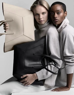 Two female models wearing cream turtleneck sweaters holding large leather tote bags against a grey studio backdrop.