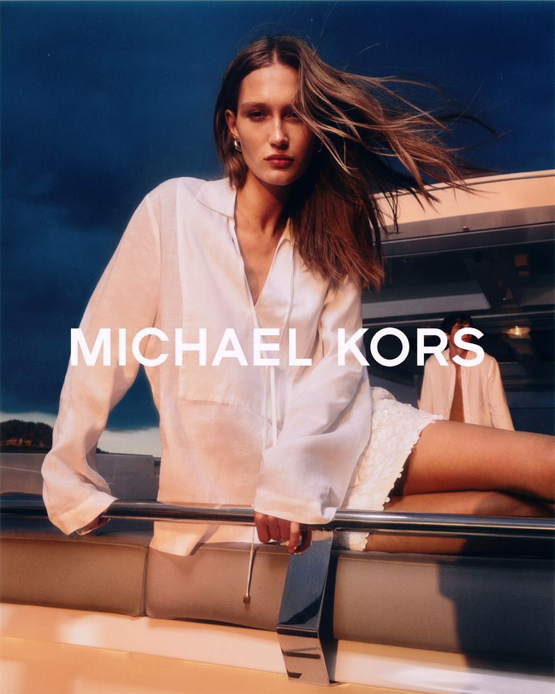 Fashion shot featuring a woman in a white shirt on a boat deck against a cloudy sky, promoting the Michael Kors brand.