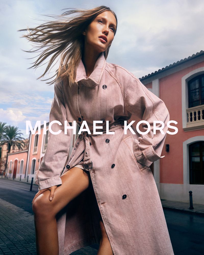 Blonde-haired woman poses in a pink trench coat in front of a blush building for a fashion advertisement with Michael Kors branding.