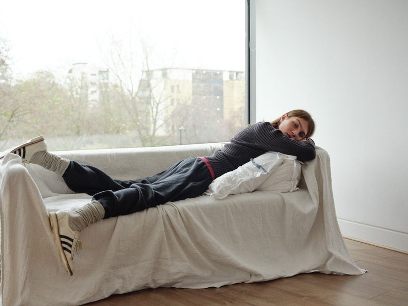 Adult woman relaxing on a cream couch in a brightly lit room with athletic shoes, grey socks, and dark sweatpants. Large window view in background.