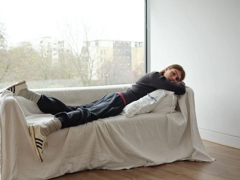 Adult woman relaxing on a cream couch in a brightly lit room with athletic shoes, grey socks, and dark sweatpants. Large window view in background.