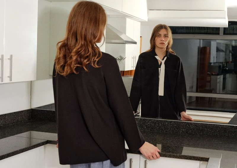 Reflection of a young adult in a black shirt with a white tie looking into a mirror, with a second blurred figure in the foreground.