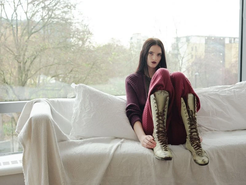 A brunette woman sits on a white couch with tall lace-up boots and maroon sweater and pants near a large window with an outdoor view.