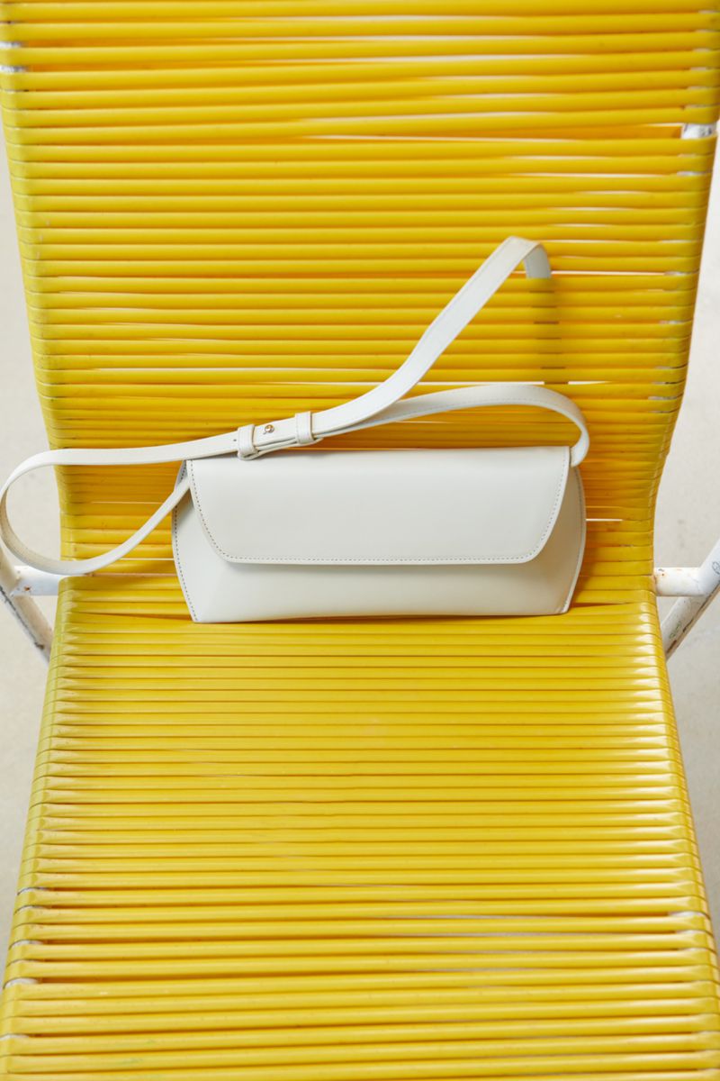 Studio shot of a white leather crossbody bag on a yellow chair. The bag has a flap closure and adjustable strap, simple design.
