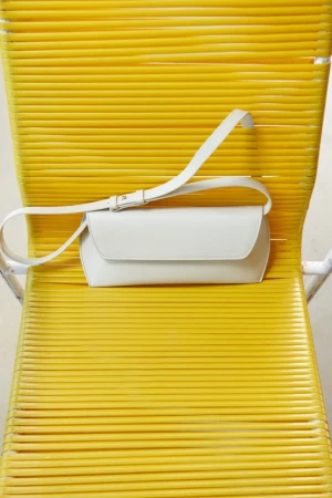 Studio shot of a white leather crossbody bag on a yellow chair. The bag has a flap closure and adjustable strap, simple design.