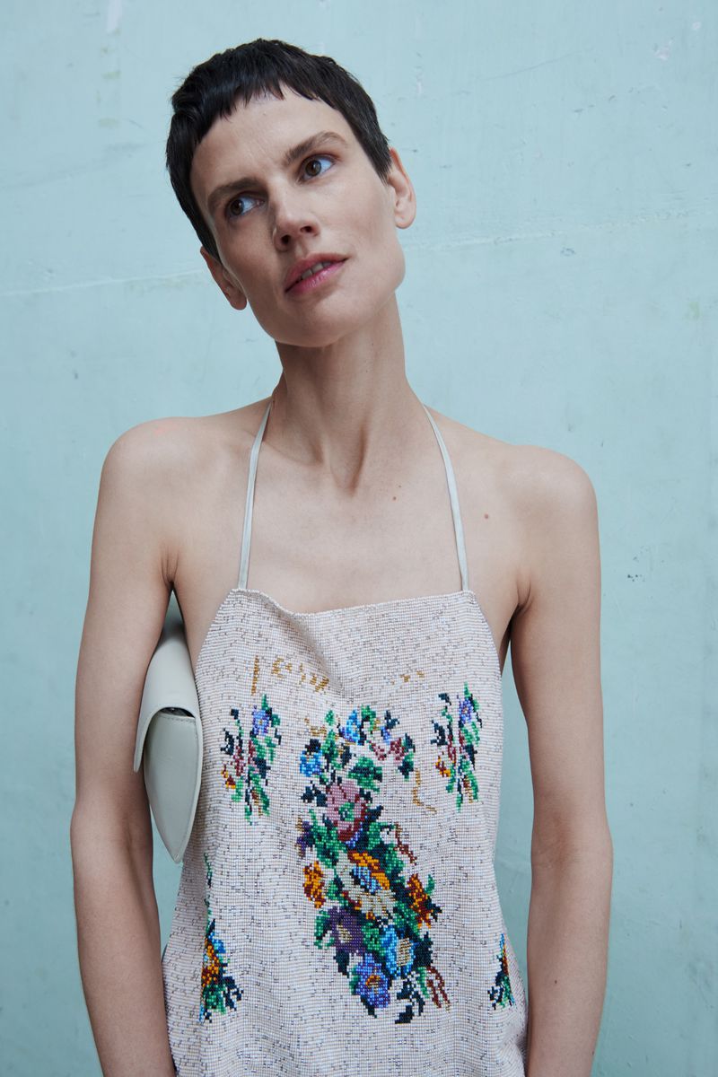Fashion shot of a woman with short brown hair wearing an embroidered dress, looking up and away from the camera in an indoor setting.