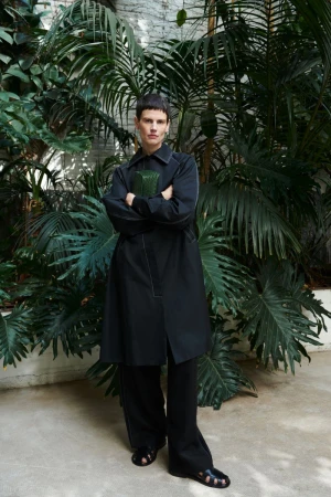 Standing model in a black trench coat, black pants and black sandals, holding a green clutch, posing in front of tropical plants.