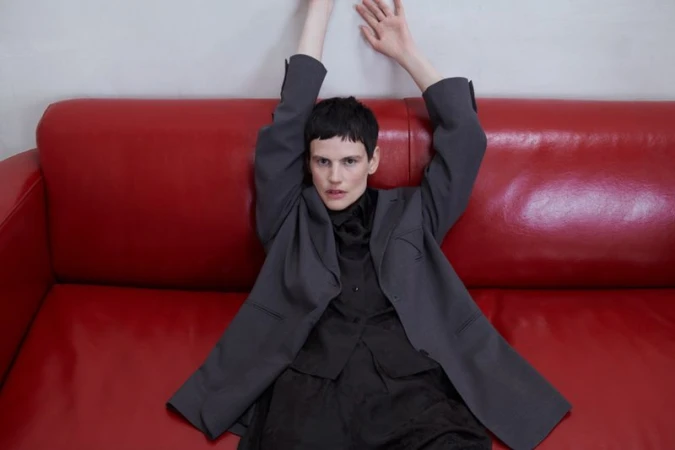 Woman in dark blazer and blouse laying back on a red couch with arms raised in a studio setting, fashion editorial composition.