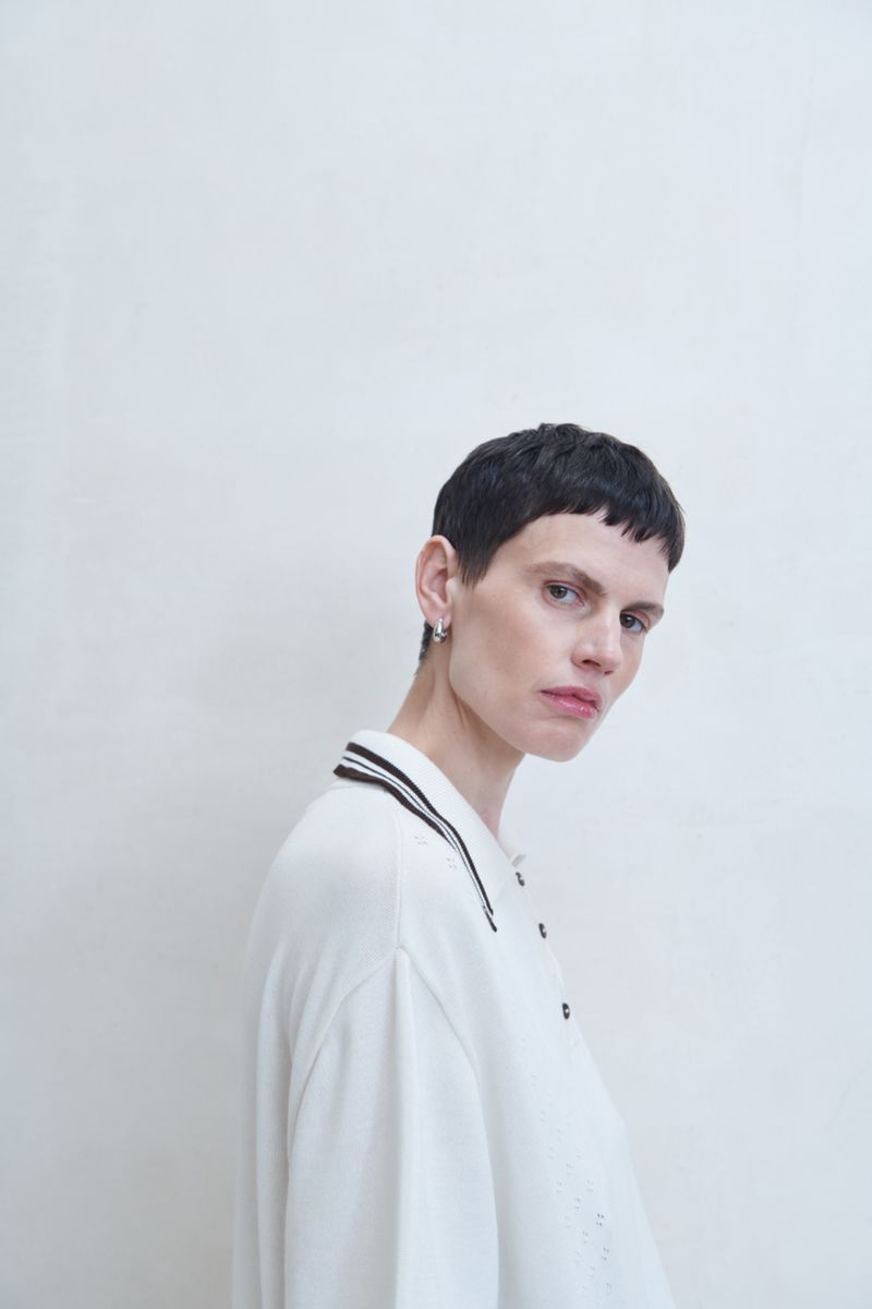 Portrait of an adult female with short dark hair, wearing a white polo shirt in front of a white wall. Studio lighting.