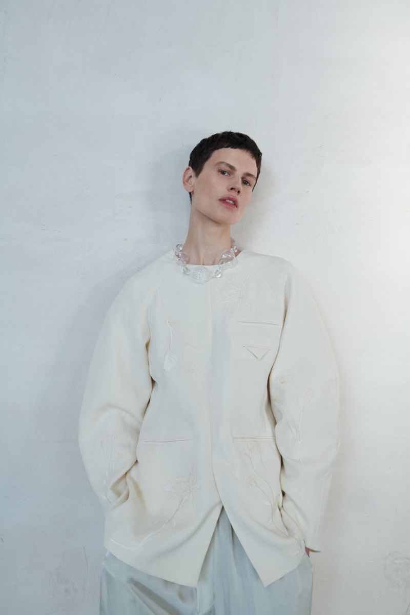 Woman with short hair wears an off-white oversized blazer and necklace against a plain white textured wall indoors, portrait orientation.
