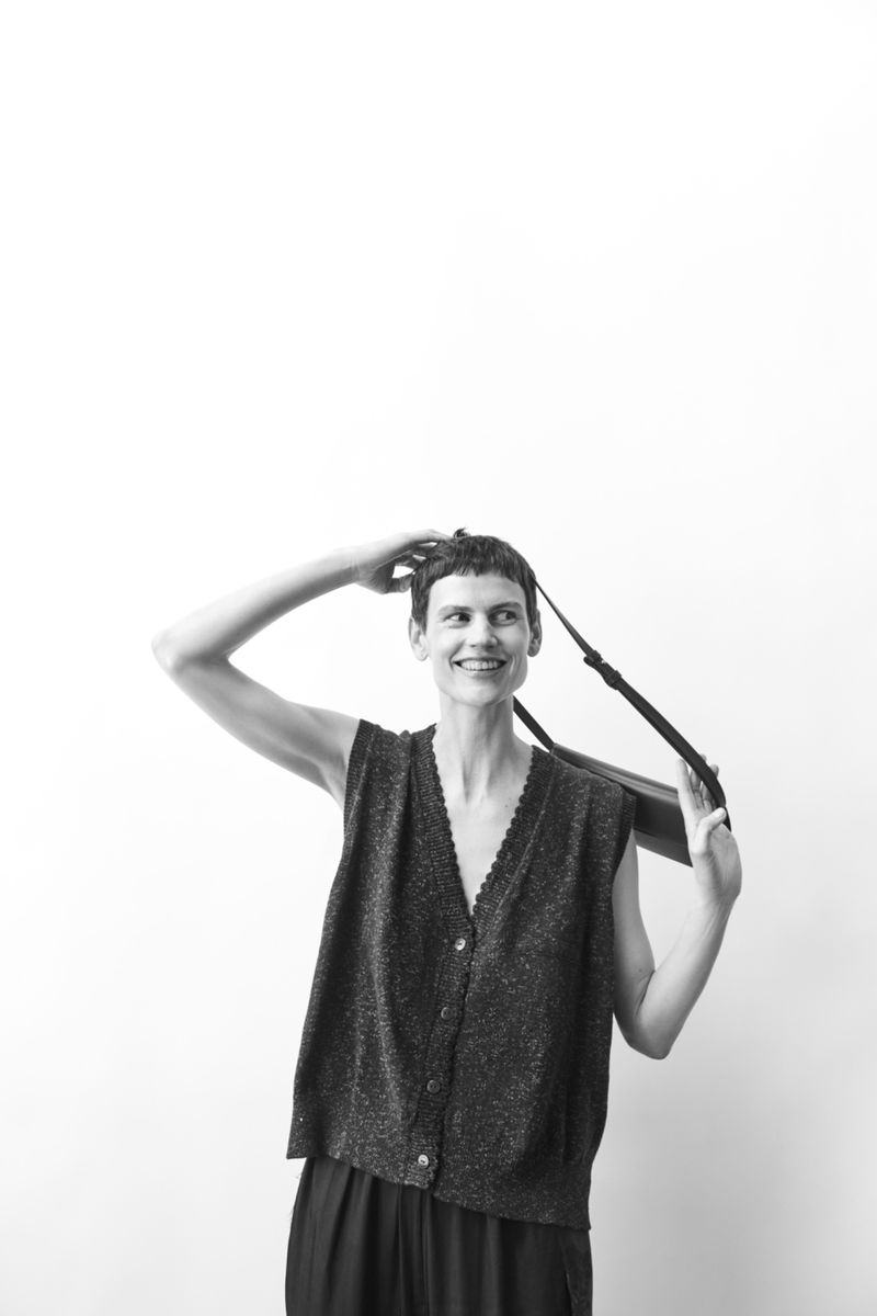 A woman poses in a sleeveless vest with a strap over her shoulder, set against a stark white backdrop in a black and white portrait.