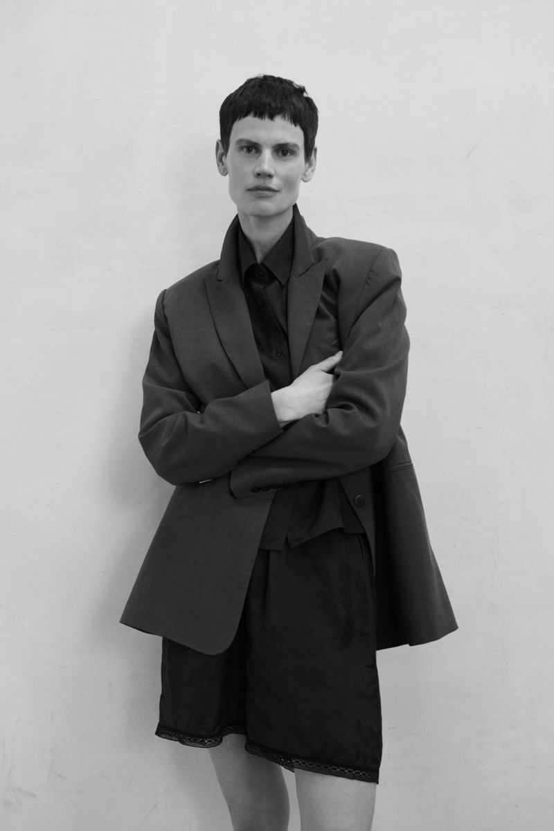 Black-and-white shot of androgynous model in oversized blazer and black shorts against a white wall, arms crossed, looking at viewer.