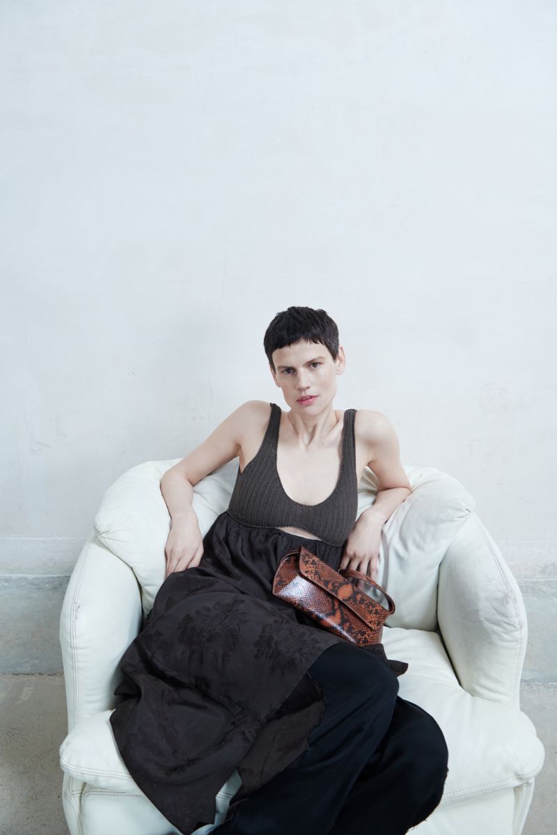 Woman with short dark hair sits in white armchair wearing a brown dress and black pants, holding a brown snakeskin purse.