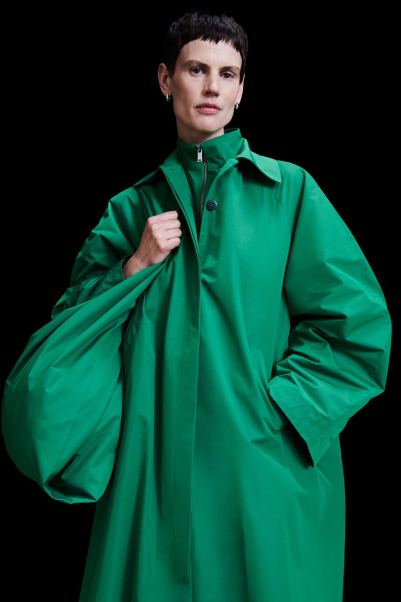 Woman in a bright green coat holding a large bag against a black background. Fashion shot with a solid, dark studio backdrop.