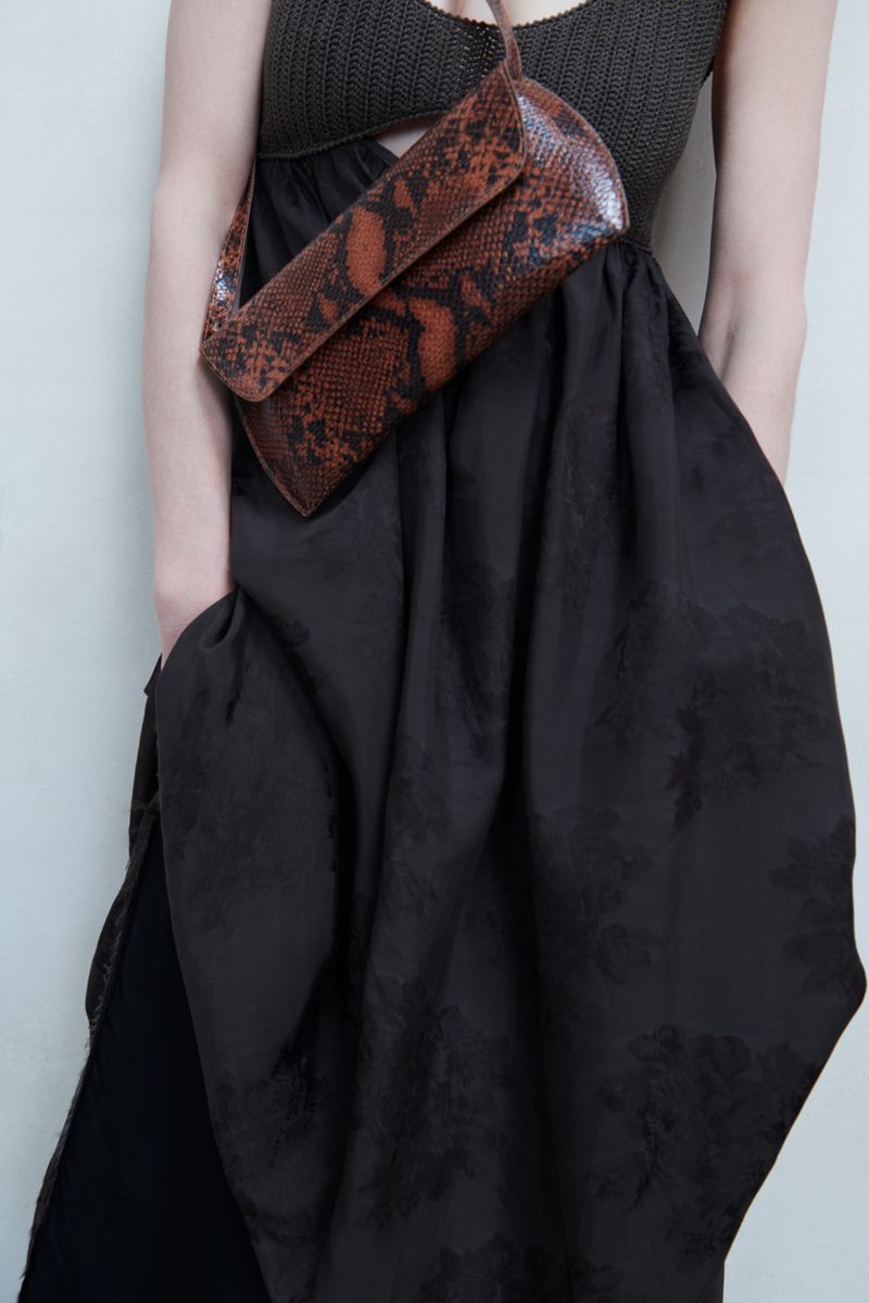 Model wearing a dark textured dress and carrying a brown snakeskin crossbody bag against a plain wall