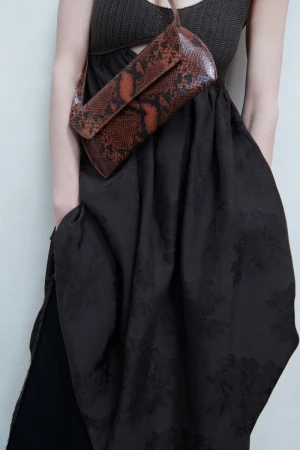 Model wearing a dark textured dress and carrying a brown snakeskin crossbody bag against a plain wall