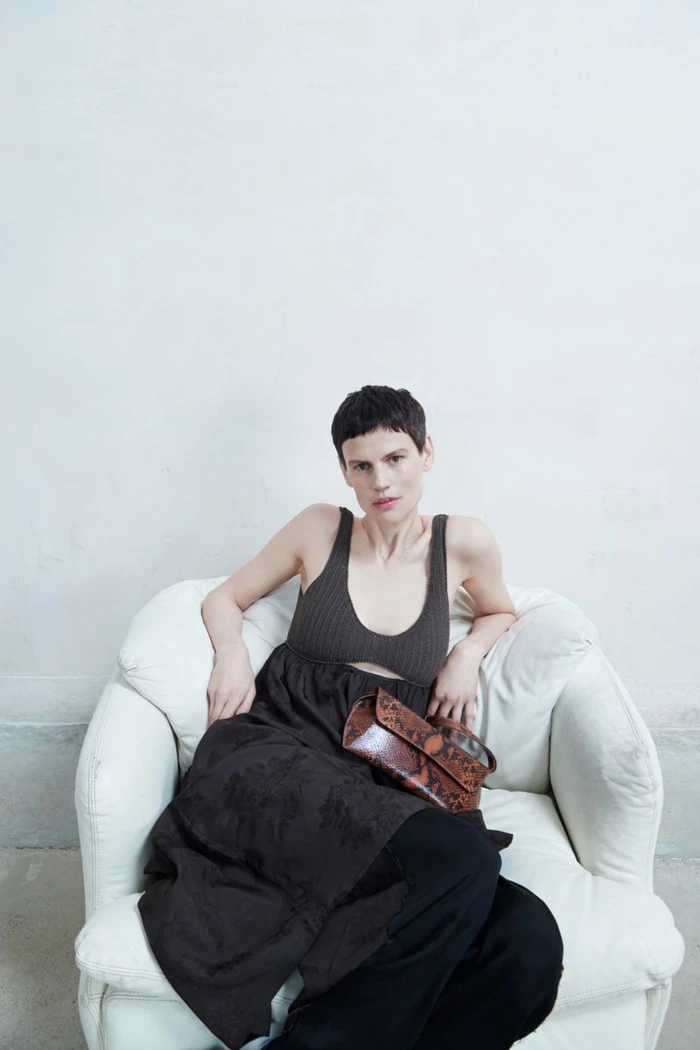 Model seated in a white armchair wearing a dark ribbed tank top and dark skirt holding a patterned handbag.
