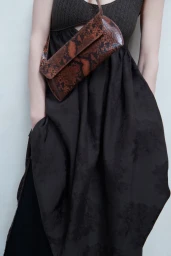 Model wearing a dark textured dress and carrying a brown snakeskin crossbody bag against a plain wall