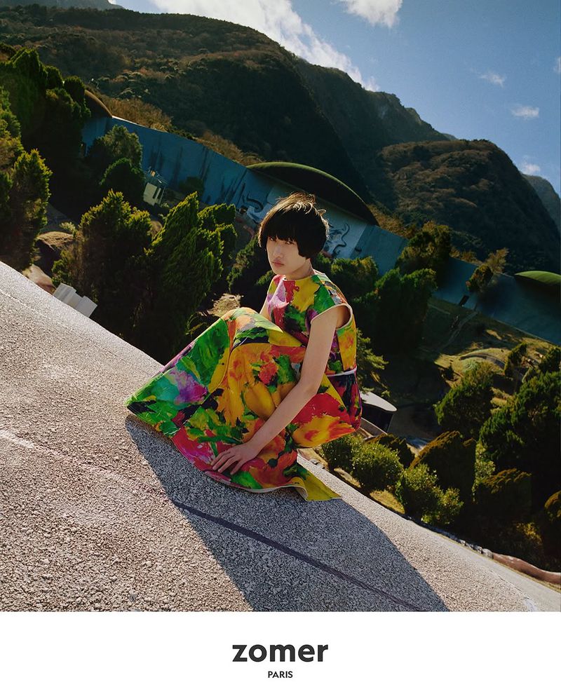 Woman in colorful floral dress squats on rooftop with mountain range background. Fashion shot with natural light and unique perspective.