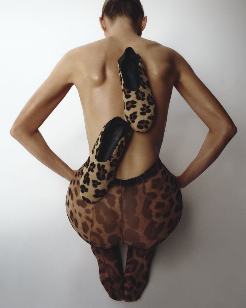 Rear view of an adult woman with fair skin wearing leopard print tights and balancing leopard print ballet flats on her back.
