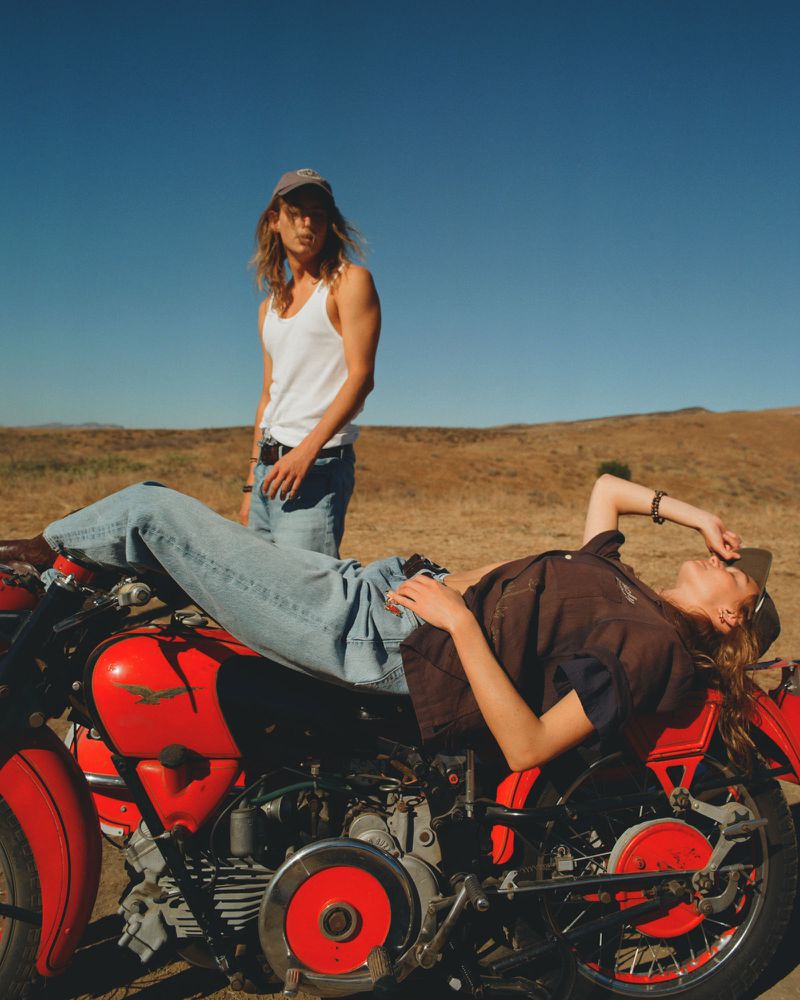 Two people pose with a vintage red motorcycle on a dirt field under a blue sky, one reclining on the bike, the other standing.