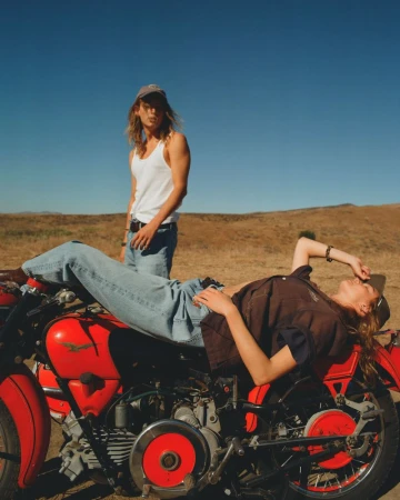 Two people pose with a vintage red motorcycle on a dirt field under a blue sky, one reclining on the bike, the other standing.