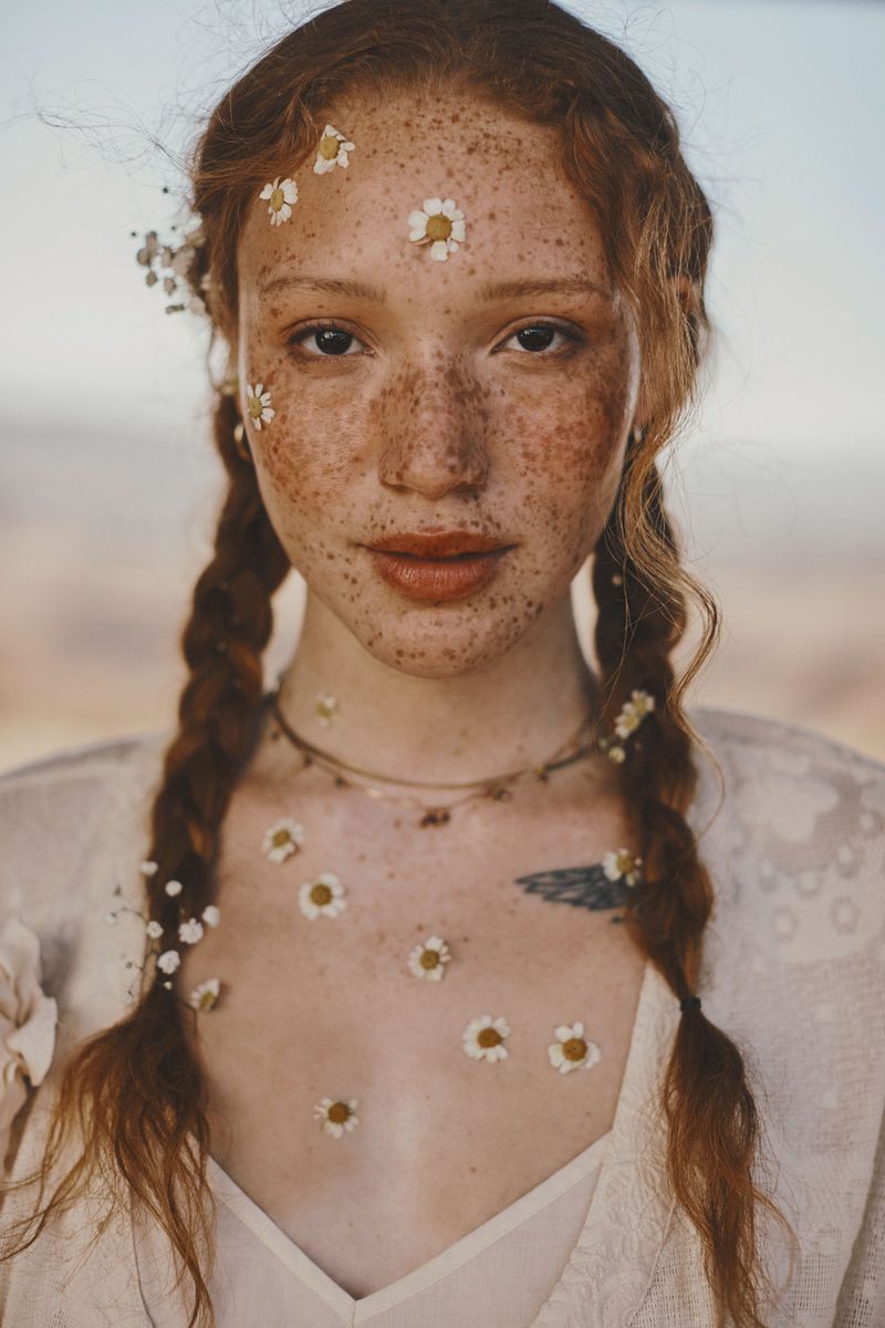 Close-up of a woman with freckles and braided red hair decorated with daisies, wearing a white top, set against a soft, blurred background.