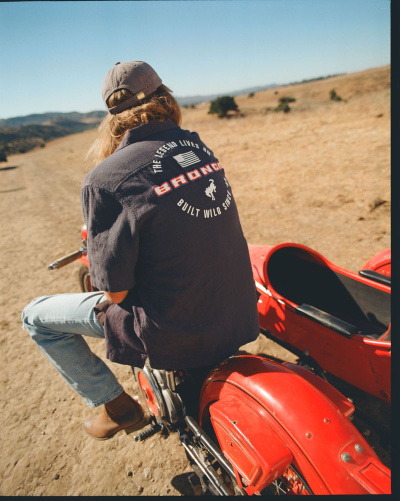 Rear view of person sitting on red motorcycle wearing cap, denim jeans, and a dark shirt with 'Bronco' logo on a sunny, rural day.