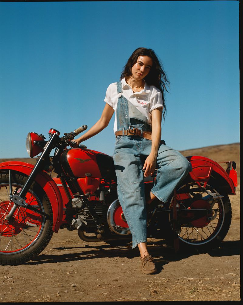 A woman in denim overalls sits on a red motorcycle in a sunny outdoor scene. The sky is clear blue and the background is muted brown.