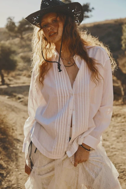Young woman in a white blouse and cowboy hat stands outside in a desert landscape with trees and a mountain in the background.