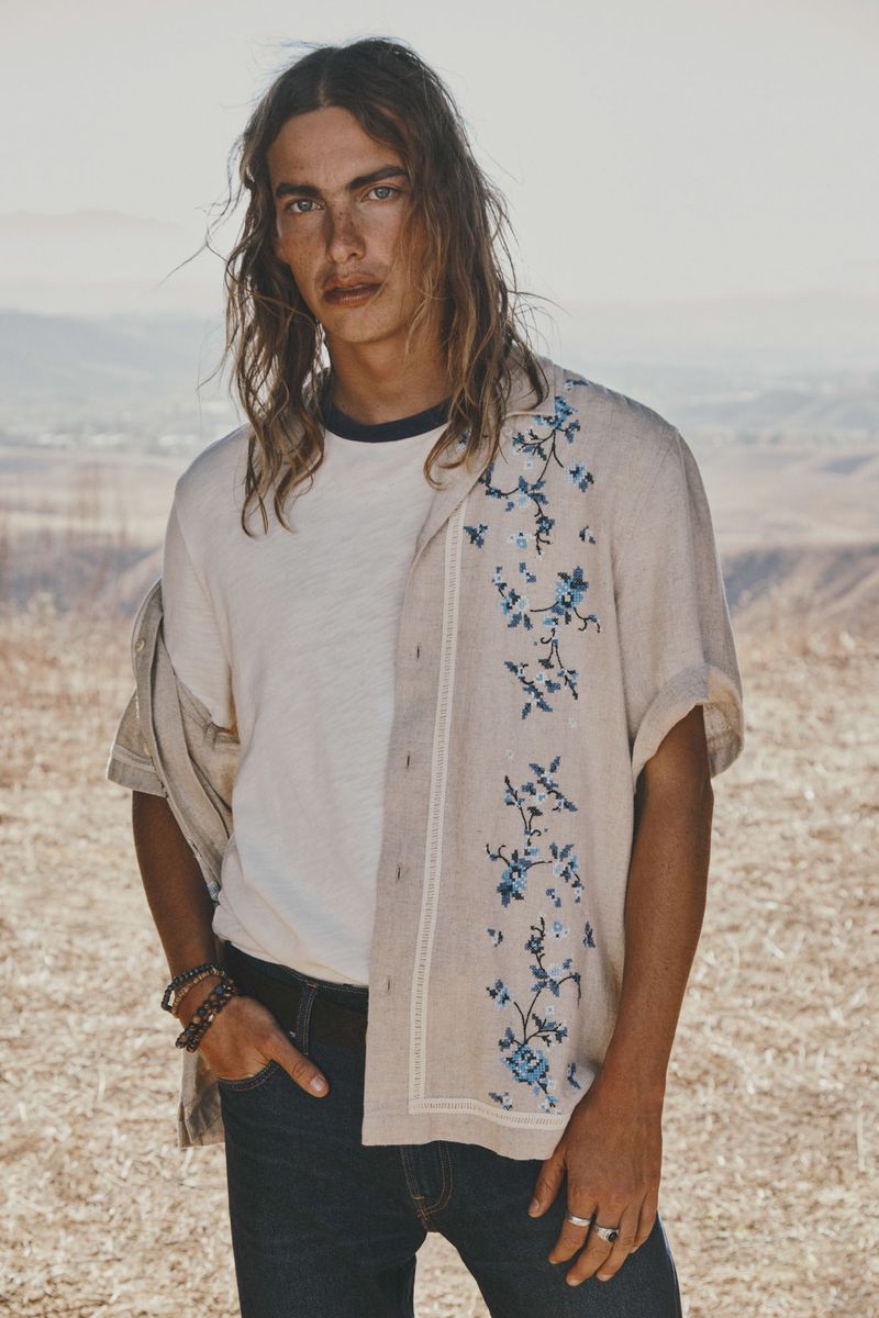 A man poses in a shirt with floral designs, wearing dark jeans, outside in a bright, earth-toned environment, looking at the camera.
