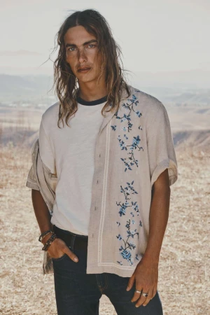 A man poses in a shirt with floral designs, wearing dark jeans, outside in a bright, earth-toned environment, looking at the camera.