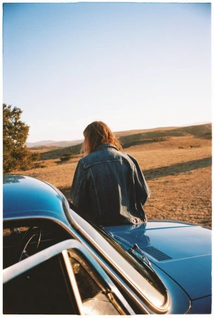 An adult wearing a denim jacket sitting on the hood of a vintage blue car, viewed from behind, with a rural landscape in the background.
