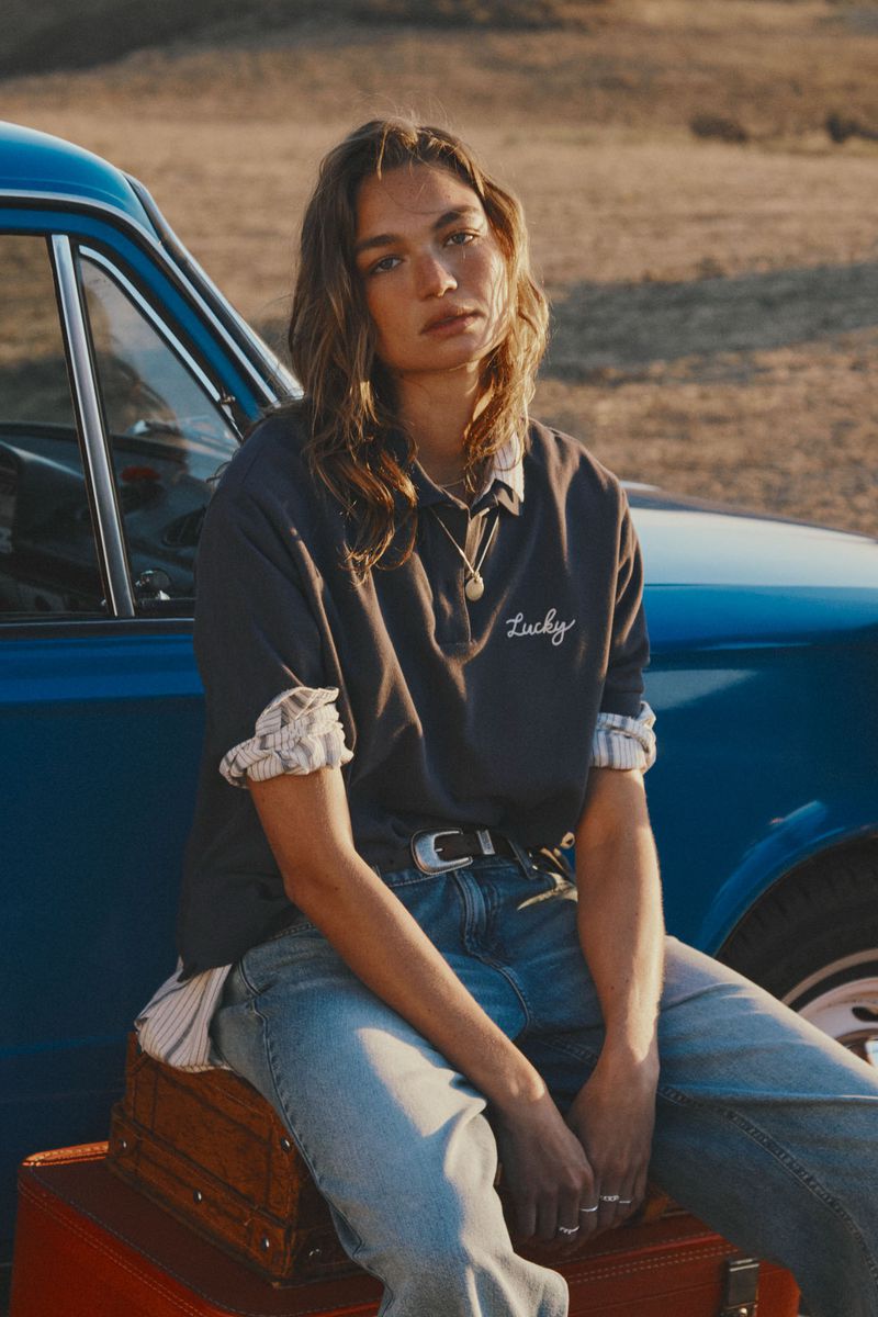 A young adult woman sits atop a brown suitcase, positioned in front of a classic blue car against a sunny, tan-colored outdoor backdrop.