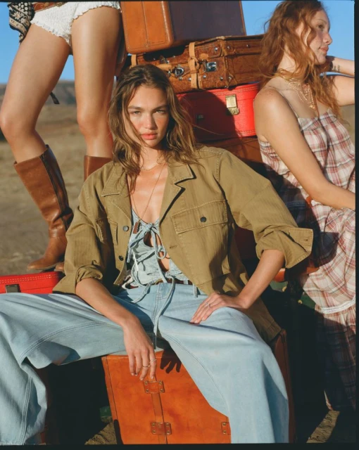 Three women pose in a desert setting, dressed in vintage-style clothing and accessorized with leather suitcases, under a clear blue sky.
