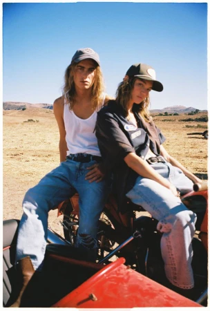 Two people wearing ballcaps pose on a motorcycle against a desert backdrop under a bright blue sky on a sunny day.