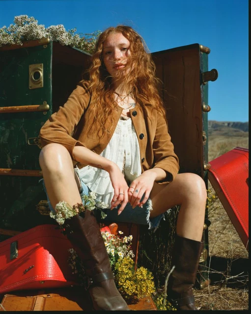 Young woman with auburn hair sits among vintage luggage, wearing a tan jacket and brown boots against a clear blue sky.