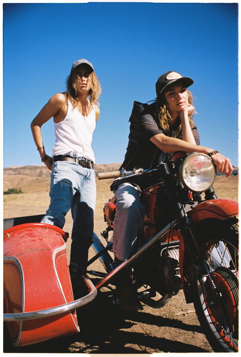 Two women pose outdoors with a vintage motorcycle and sidecar, set against a clear blue sky and arid, hilly landscape.