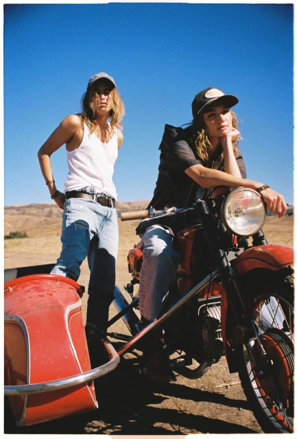 Two women pose outdoors with a vintage motorcycle and sidecar, set against a clear blue sky and arid, hilly landscape.