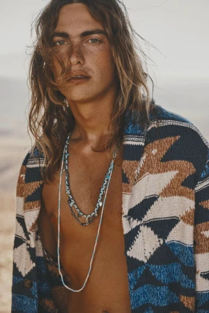 Close-up of a young adult male model wearing a patterned cardigan and necklaces, posing outdoors in a bright, sunlit environment.