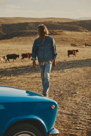 Person in denim jacket and jeans walks past a blue car on a ranch with cows and golden hills in the background during daytime.