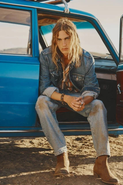 Man with long blond hair wearing denim jacket and jeans sitting in a blue car on a sunny day in the desert.
