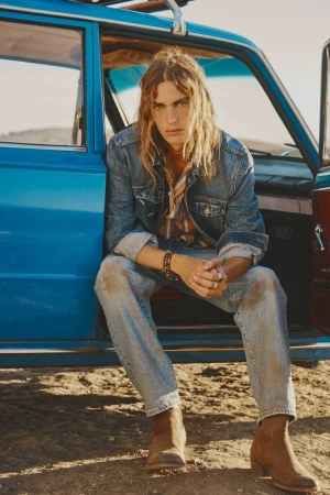 Man with long blond hair wearing denim jacket and jeans sitting in a blue car on a sunny day in the desert.