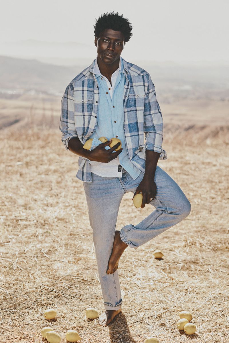 An African American man balances barefoot with a pile of lemons in hand on a sunny, grassy field, wearing a blue plaid shirt and jeans.
