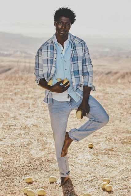 An African American man balances barefoot with a pile of lemons in hand on a sunny, grassy field, wearing a blue plaid shirt and jeans.