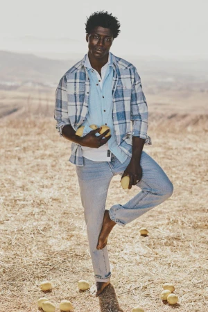 An African American man balances barefoot with a pile of lemons in hand on a sunny, grassy field, wearing a blue plaid shirt and jeans.