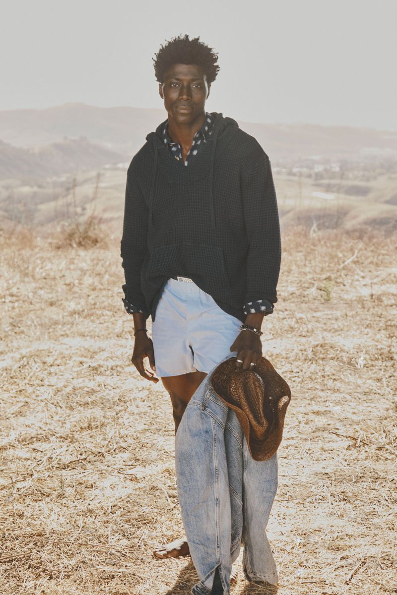 Man in hoodie stands in a sunlit field with a jacket and hat. Hilly landscape fades into a bright sky. Casual fashion shot outdoors.