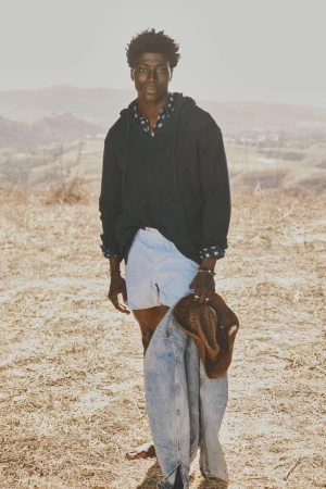 Man in hoodie stands in a sunlit field with a jacket and hat. Hilly landscape fades into a bright sky. Casual fashion shot outdoors.