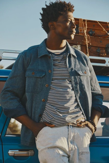 Man in denim jacket and striped t-shirt stands beside a blue car with luggage on top, enjoying natural outdoor lighting.