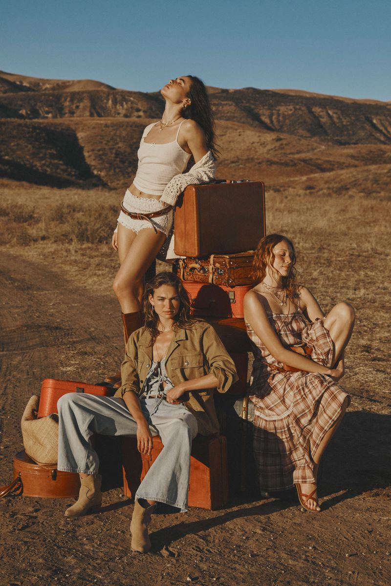 Three women in casual fashion, posing near vintage luggage in a dry, hilly landscape with a clear blue sky in an outdoor fashion editorial.